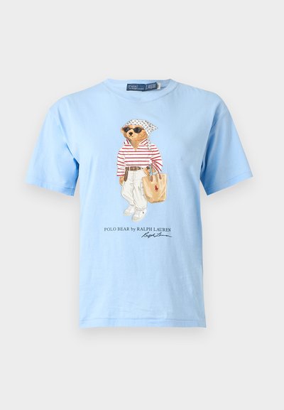 Ανοιχτό μπλε T-shirt με μια αρκούδα που φορά γυαλιά ηλίου, ριγέ πουκάμισο, λευκό παντελόνι και κρατά μια μπεζ τσάντα tote, με το κείμενο «Polo Bear by Ralph Lauren» από κάτω.