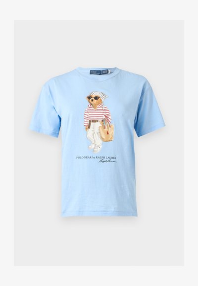 Ανοιχτό μπλε T-shirt με μια αρκούδα που φορά γυαλιά ηλίου, ριγέ πουκάμισο, λευκό παντελόνι και κρατά μια μπεζ τσάντα tote, με το κείμενο «Polo Bear by Ralph Lauren» από κάτω.