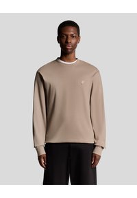 Beige sweatshirt med lange ærmer, rund halsudskæring og broderet logo på brystet. Glat tekstur, afslappet pasform, parret med sorte bukser.