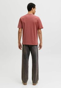 Hombre con cabello rizado corto vestido con una camiseta roja polvorienta y jeans negros sueltos y desgastados, de pie mirando hacia el lado opuesto, contra un fondo claro y liso.