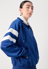 Jaqueta azul com fecho éclair, apresentando um colarinho branco e detalhes listrados nas mangas, com punhos canelados e bolsos laterais. Textura suave.