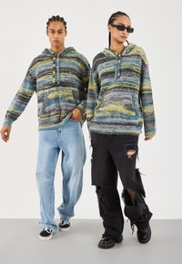 Knitane kapuce s horizontalnimi črtami v modri, rumeni in sivi barvi. En model ima na sebi svetlo modre džins hlače, drugi pa uničene črne hlače in škornje.