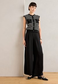 Haut sans manches à rayures noires et blanches avec fermeture à boutons, associé à un pantalon large noir et des chaussures plates noires. Texture plate et coupe décontractée.