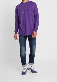 Lila långärmad skjorta med en guldig logotyp på bröstet, tillsammans med blå jeans och marinblå sneakers med vita detaljer. Avslappnad passform och material.