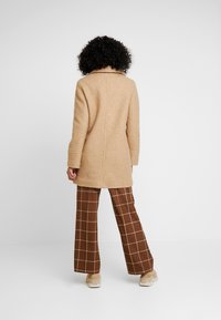 Beige textured coat with a slightly loose fit, featuring a collar and long sleeves. Paired with brown plaid wide-leg trousers and light footwear.