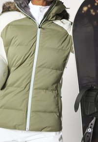 Giacca da sci imbottita di colore verde oliva con cappuccio foderato in pelliccia, dotata di chiusura con zip e pannelli bianchi a contrasto, tenuta accanto a uno snowboard.