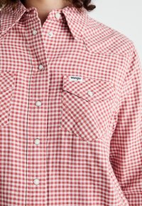 Camicia in tessuto a quadri rossi e bianchi con colletto button-down, due tasche sul petto e logo Wrangler. Il tessuto sembra leggero e di consistenza testurizzata.
