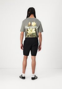 Szary bawełniany t-shirt z kwiatowym wzorem oraz słowem "Harmonia" w białym kolorze. W połączeniu z czarnymi szortami, model nosi czarne buty.