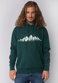 Man met lang haar die een groene hoodie draagt met een witte berg- en bosafbeelding, staand tegen een effen lichte achtergrond.