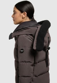 Giacca puffa marrone con chiusura zip, colletto foderato in pelliccia e logo Khujo sulla manica. Presenta un motivo trapuntato e cinturini regolabili.