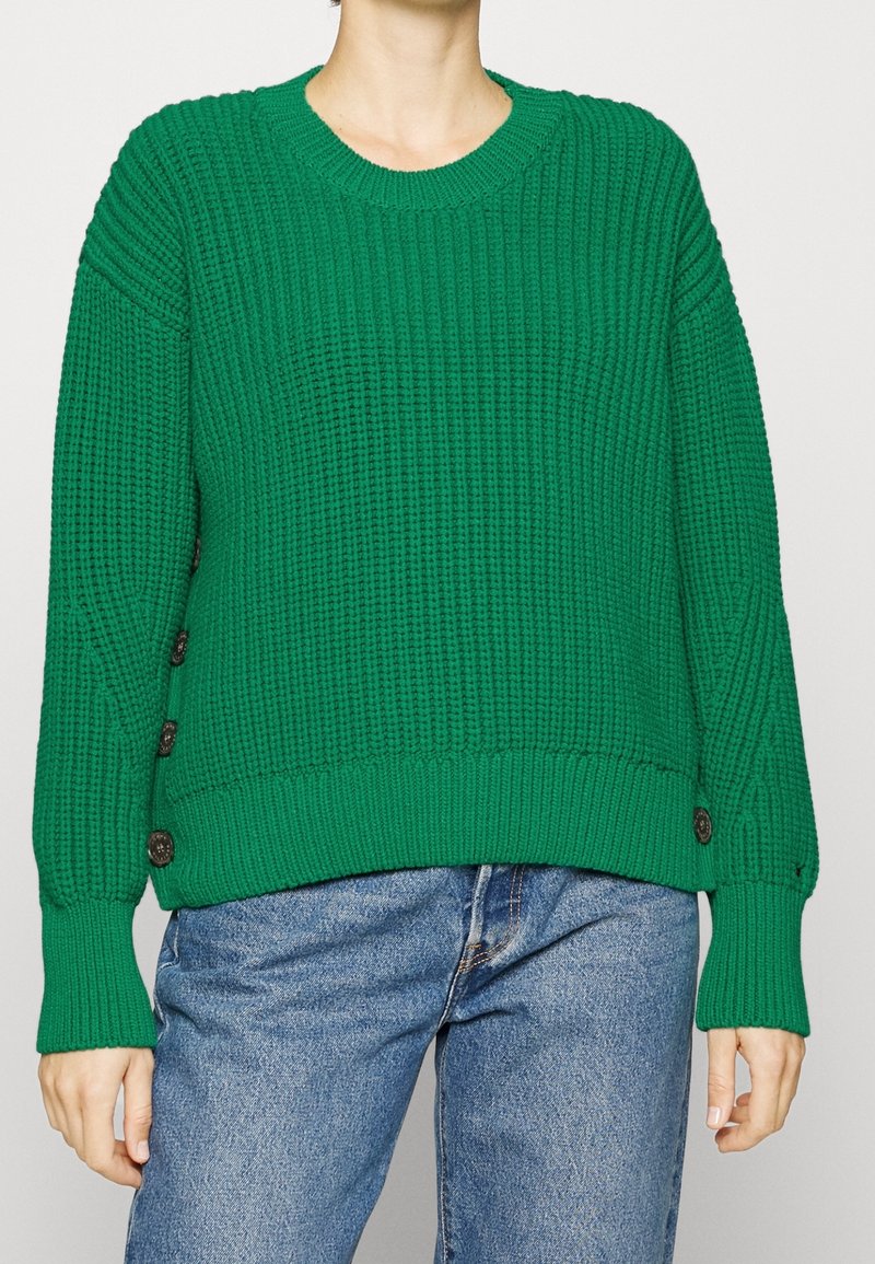 Pull en tricot vert avec une texture côtelée, un col rond et de gros boutons sur les côtés. Coupe décontractée avec des manches longues et un ourlet court.