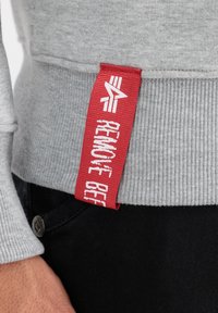 Szary materiał bluzy z prążkowaną fakturą, zawierający czerwoną metkę z napisem "REMOTE BEER". Czarne spodnie widoczne na dole obrazu.