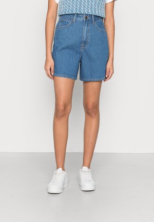Persoon die middenlange blauwe spijkershorts, een lichtblauw top met patroon en witte sneakers draagt, staand tegen een effen lichte achtergrond.
