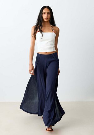 Femme marchant avec un crop top blanc sans manches avec des bretelles en perles roses, associé à un pantalon large bleu marine avec panneaux latéraux.