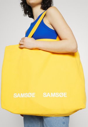 Osoba w niebieskim topie bez rękawów trzyma dużą żółtą torbę na ramię z białym napisem "SAMSØE SAMSØE", ubrana w jasnoniebieskie jeansy na białym tle.