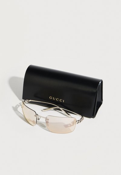 Lunettes de soleil rectangulaires sans monture avec des verres légèrement teintés et des branches argentées, exposées devant un étui noir de marque Gucci.