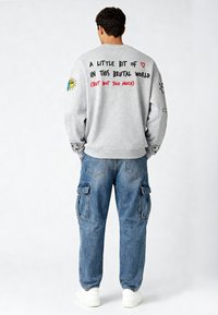 Felpa grigia con testo colorato sulla schiena e design ricamati sulle maniche, abbinata a jeans cargo blu e sneakers bianche.