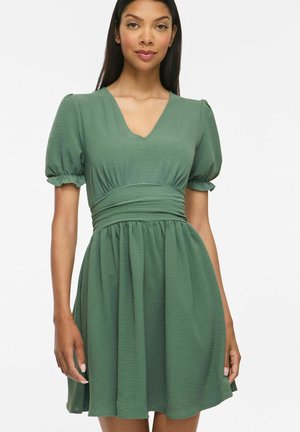 Femme aux longs cheveux foncés portant une robe verte à manches courtes avec un profond décolleté en V et une taille froncée, debout sur un fond blanc.