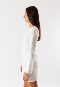 Abito bianco a maniche lunghe con scollo rotondo e design aderente, realizzato in tessuto morbido e con taglio al ginocchio, presentato su uno sfondo neutro.