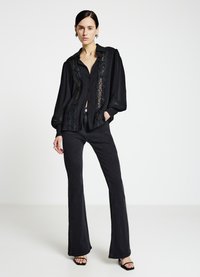 Schwarzes Hemd mit Spitzenbesatz und Knopfleiste, kombiniert mit eng anliegenden schwarzen Schlaghosen und schwarzen Sandalen mit Absatz, vor neutralem Hintergrund.