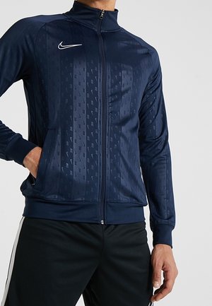 Homme portant une veste zippée Nike bleu marine avec des motifs verticaux subtils et un logo Nike blanc sur la poitrine, associée à un pantalon noir.