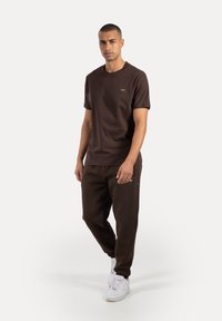 Czekoladowy brązowy t-shirt i pasujące dresy, wykonane z miękkiego materiału. Zawiera małą metkę na koszulce i kieszeń w spodniach.