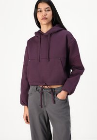 Felpa corta viola con cappuccio, cordoncino sull'orlo e logo testurizzato sulla manica sinistra. Abbinata a pantaloni grigi.