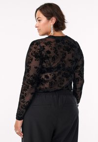 Sort gennemsigtig langærmet bluse med blomstermotiver i blondestof, rund halsudskæring og tætsiddende manchetter, sat sammen med højtaljede sorte bukser.