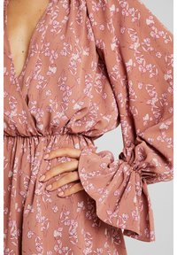 La main d'une femme repose sur la taille, elle porte une robe rose poussiére avec un imprimé floral blanc et des manches bouffantes froncées avec des poignets à volants.
