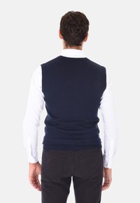 Maglione senza maniche di colore blu navy realizzato in tessuto a maglia, con scollatura rotonda e orlo a coste, indossato sopra una camicia bianca a maniche lunghe.