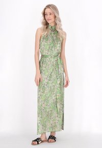 Rochie maxi cu model paisley verde, fără mâneci, cu guler înalt, talie cu curea, material fluid, purtată cu sandale negre cu curele.