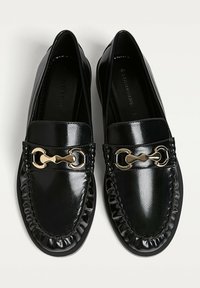 Černé kožené mokasíny s lesklým povrchem, s akcentem v podobě zlatého řetízku a staženým okrajem. Hladká textura a zaoblený design špičky.