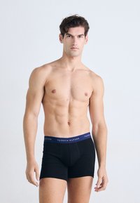 Sorte bomulls-boxershorts med en marineblå livrem som har "TOMMY HILFIGER" i hvit tekst. Glatt tekstur, tettsittende design, midt på låret lengde.