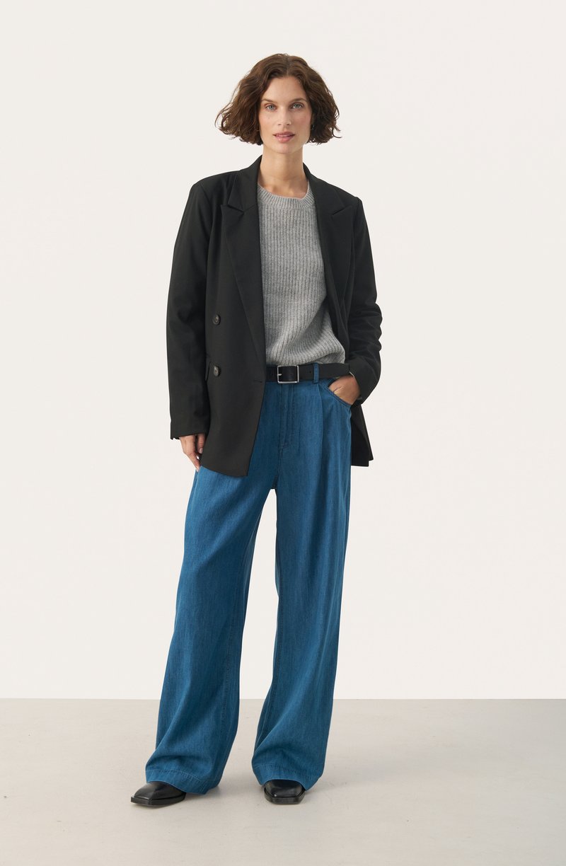 Schwarze doppelt geknöpfte Blazer, hellgrauer gerippter Pullover, weite blaue Jeanshosen, schwarzer Gürtel und schwarze Schuhe vor einem neutralen Hintergrund.