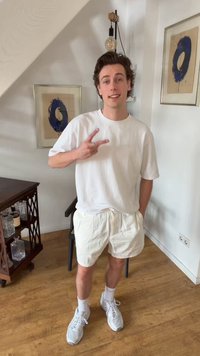 Joven vestido con camiseta blanca, pantalones cortos y zapatillas deportivas haciendo el signo de la paz, de pie en una habitación con suelo de madera y arte abstracto en las paredes.
