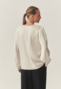 Kremna bluza z dolgimi, razširjenimi rokavi in zapenjanimi manšetami, sokroglim izrezom in gladko teksturo, kombinirana s črnimi hlačami.