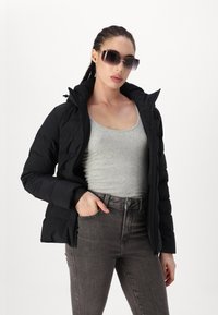 Chaqueta acolchada negra con un cuello alto, usada sobre una camiseta sin mangas gris, combinada con jeans gris oscuro. Incluye bolsillos frontales y cierre de cremallera.
