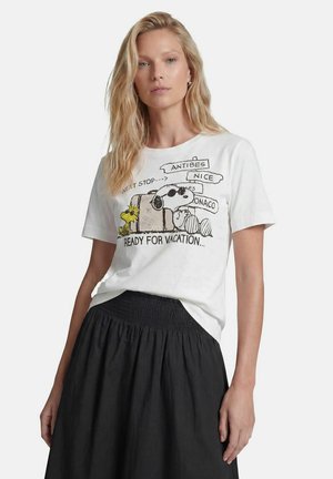 Blonde Frau trägt ein weißes T-Shirt mit Snoopy- und Woodstock-Cartoon und einen schwarzen Rock, steht vor einem schlichten hellen Hintergrund.