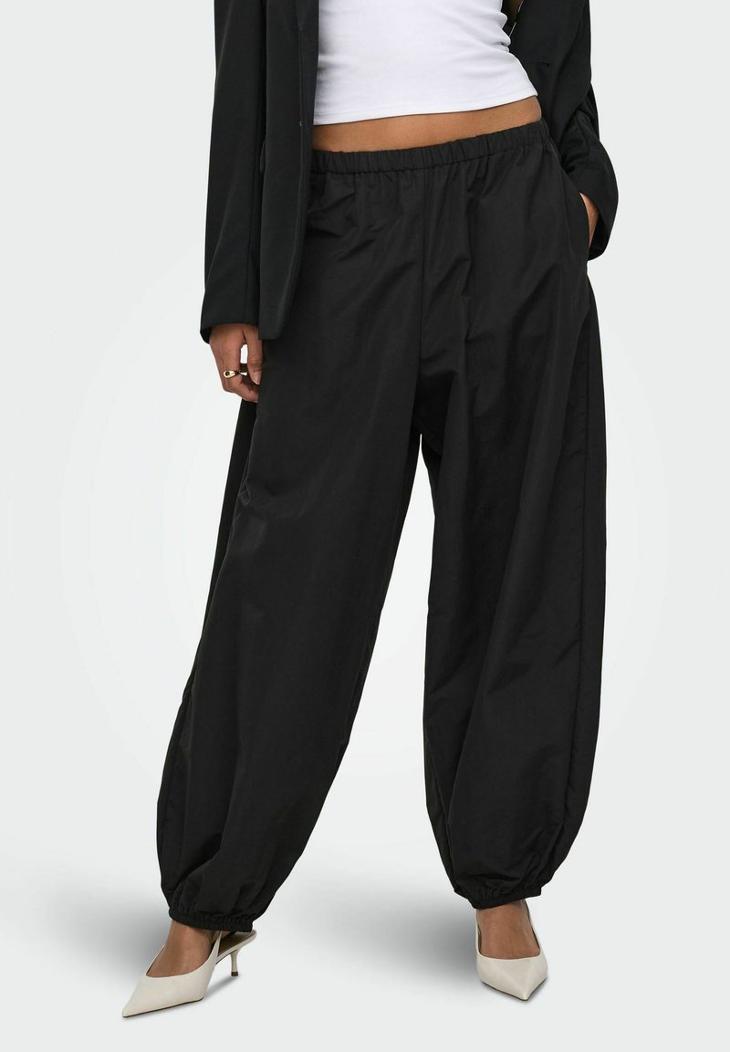 Femme portant un pantalon ample noir à taille élastique avec des poignets élastiques, un top blanc court, un blazer noir et des talons blancs pointus.