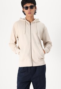 Beige színű, cipzáros kapucnis jumper, puha anyagból készült, állítható kapucnival, elülső zsebekkel és laza fazonnal, sötétkék nadrággal párosítva.