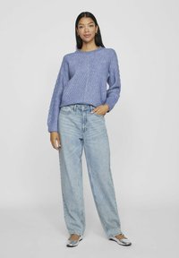 Ljuslila stickad tröja med ett texturerat mönster, kombinerad med ljusblå högmidjade jeans och silverfärgade ballerina skor. Modellen poserar med en hand på höften.