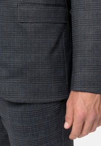 Grau kariertes Anzug mit feinem Überprüfungs-Muster in dunkleren Farbtönen. Der Stoff ist strukturiert, und eine Tasche ist auf der Jacke sichtbar.