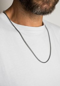 Colier din metal cu un finisaj neted, argintiu, având un design răsucit, purtat peste un tricou polo alb simplu.