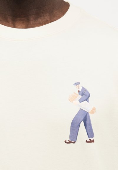 Άνδρας ντυμένος με vintage μπλε ρούχο, κρατώντας μια μεγάλη μπαγκέτα τυλιγμένη σε λευκό χαρτί στην περιοχή της τσέπης ενός λευκού μπλουζάκι.