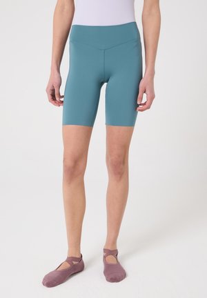 Persona con pantalones cortos de ciclismo color verde azulado hasta la mitad del muslo y calcetines estilo ballet color malva, de pie contra un fondo blanco liso.