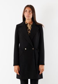 Cappotto nero doppiopetto con grandi rever, due tasche frontali, dettagli in metallo dorato, vestibilità su misura e tessuto morbido.