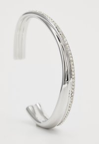 Silver cuff-armband med en slät metallfinish och en rad små klara kristaller längs en kant. Krökt design med öppna ändar.