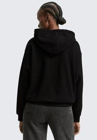 Schwarzer Kapuzenpullover mit lockerem Schnitt, gerippten Bündchen und Saum. Die Rückansicht zeigt einen geflochtenen Haarknoten und graue hochgeschnittene Jeans.