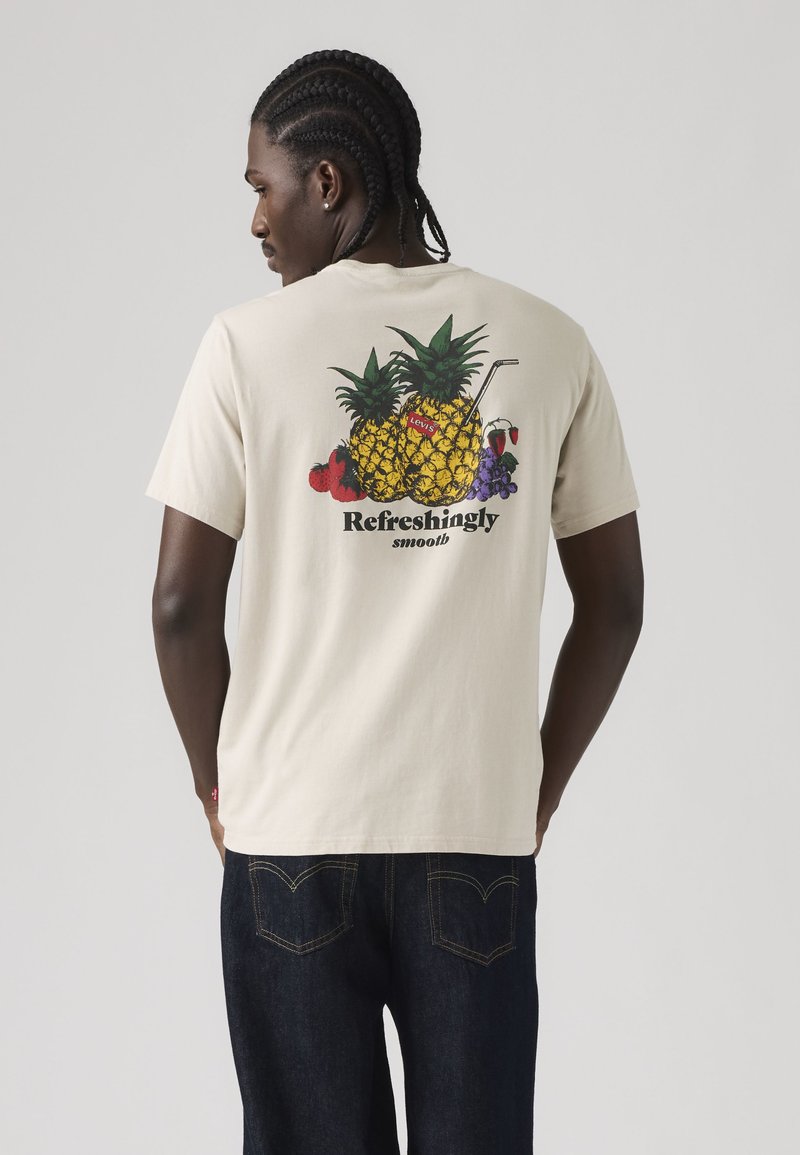 Hombre con camiseta beige con gráfico colorido de piña, fresas y uvas y texto "Refrescante y suave" en la espalda.