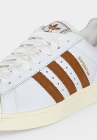 Λευκό αθλητικό παπούτσι Adidas Superstar με καφέ ρίγες, λευκά κορδόνια και χρυσό κείμενο "SUPERSTAR" στο πλάι.
